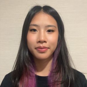 Portrait of a young woman with long black hair tinted purple at the ends, wearing a black top, neutral background, subtle smile.